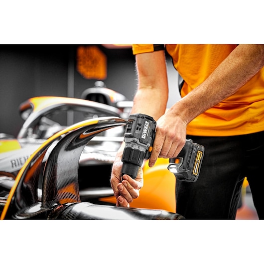 A person using a DEWALT cordless power drill (model DCD85ME2GT) to work on a Formula 1 race car, focusing on the car's aerodynamic components.