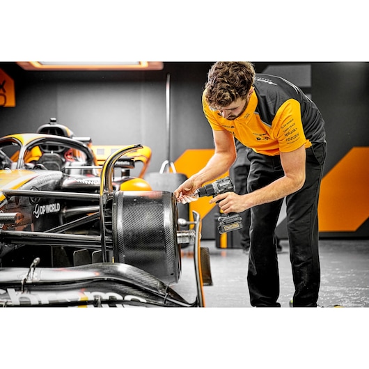 A person in a yellow and black uniform uses a DEWALT cordless drill on the wheel of a Formula 1 race car in a garage.