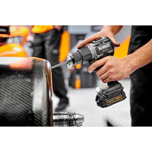 A close-up of a person using a DEWALT cordless drill to drill into a carbon fiber surface in a workshop setting. The DEWALT branding is visible on the drill.