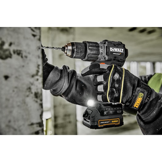A person wearing protective gloves is using a DEWALT cordless drill to drill into a concrete wall. The drill has a visible LED light and DEWALT branding. The scene takes place in an unfinished building or construction site.