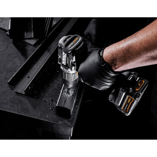 A close-up of a person wearing a black glove using a DEWALT cordless drill to drill into a metal rectangular tube. Metal shavings are visible on the surface.