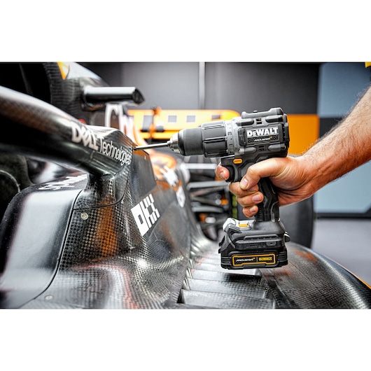 A person using a DEWALT cordless drill on the carbon fiber surface of a racing car. The drill is black and yellow, with the model DCD85ME2GT visible. The DEWALT logo is prominent on the drill.