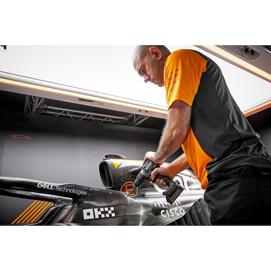 A person in a black and orange shirt using a DEWALT power drill (model DCD85ME2GT) to work on a black race car inside a garage. The car has various sponsor logos visible.