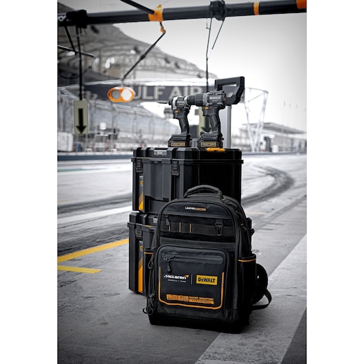 A set of DEWALT tools, including two cordless power drills, a black and yellow tool backpack, and two sturdy tool storage boxes, are arranged on the ground near a racetrack. The backpack is a limited edition with McLaren branding. The background features a pit lane and racetrack barriers.