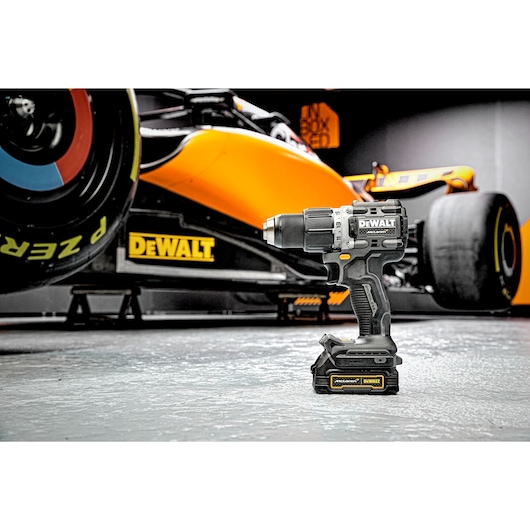 A DEWALT cordless drill (model DCD85ME2GT) is standing on a garage floor in front of an orange racing car with the DEWALT logo visible on both products.