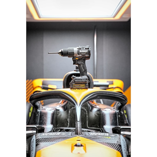 A DEWALT cordless drill model DCD85ME2GT is placed on the front section of an orange and black racing car cockpit, with the car's detailed dashboard and protective frame visible.