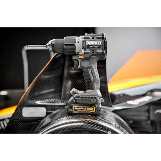 A DEWALT cordless drill, model DCD85ME2GT, is standing upright on a carbon fiber automotive part. The drill is black with yellow accents and has visible branding on the side and battery.