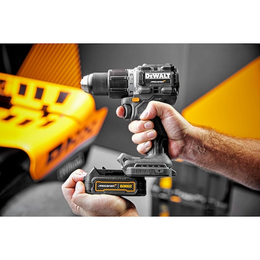 A close-up of two hands holding a DEWALT cordless power drill, with one hand inserting a removable battery pack labeled 'McLaren DEWALT'. The background features orange and black elements.