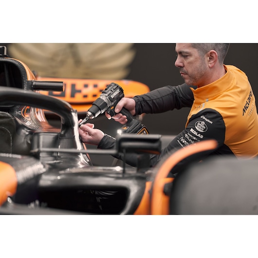 Person uses DEWALT drill on a race car part, wearing orange and black team jacket.