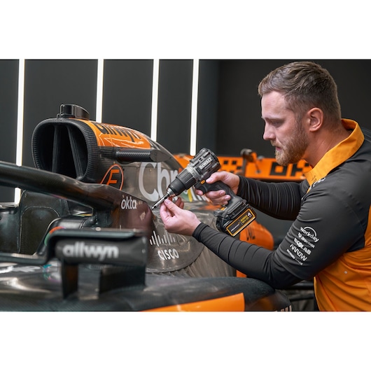 A person uses a DEWALT drill on an orange racing car, working near the cockpit area.