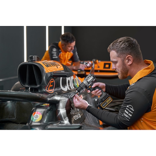 Person uses a DEWALT drill on a race car in a workshop. Faces are blurred. DEWALT branding is visible.