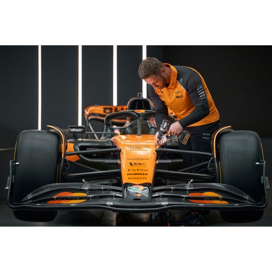 Person with blurred face uses DEWALT tool on orange race car in a modern workshop setting.