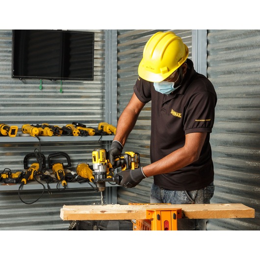 Worker using DEWALT DCD996NT drill on wood, wearing safety gear in workshop with other DEWALT tools in background.
