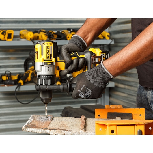 A person uses a DEWALT cordless drill to drill into metal, wearing gloves. Tools are visible on shelves nearby.