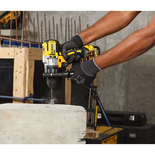Person uses a DEWALT cordless drill to bore into concrete at a construction site, wearing black gloves.