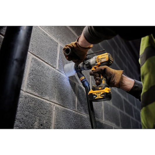 A person wearing work gloves uses a DEWALT cordless drill to bore a hole into a concrete block wall, with a dust extraction accessory attached for cleaner operation.