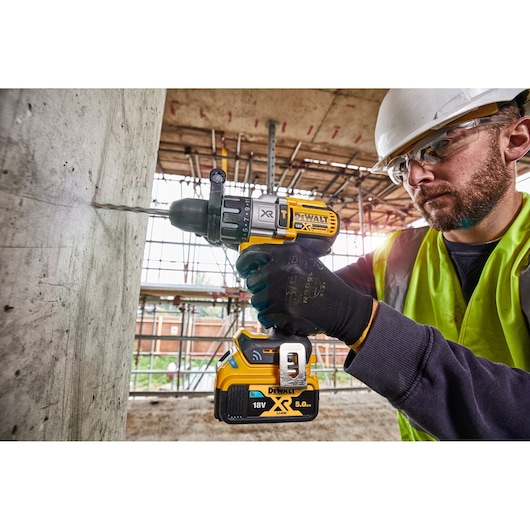 A person wearing safety gear uses a DEWALT DCD997NT 18V XR cordless drill to drill into a concrete wall at a construction site.
