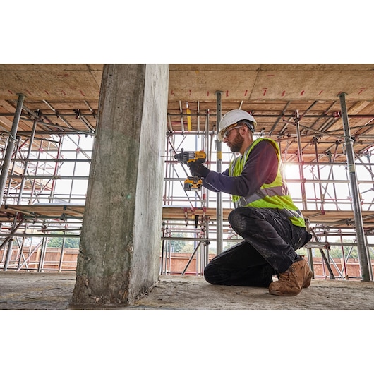 A worker in a safety helmet and high-visibility vest kneels while using a DEWALT power drill to work on a concrete pillar at a construction site with scaffolding.