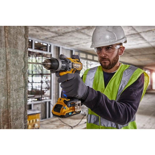 A person wearing a safety helmet, high-visibility vest, and gloves is using a DEWALT DCD997NT cordless drill to drill into a concrete wall at a construction site.