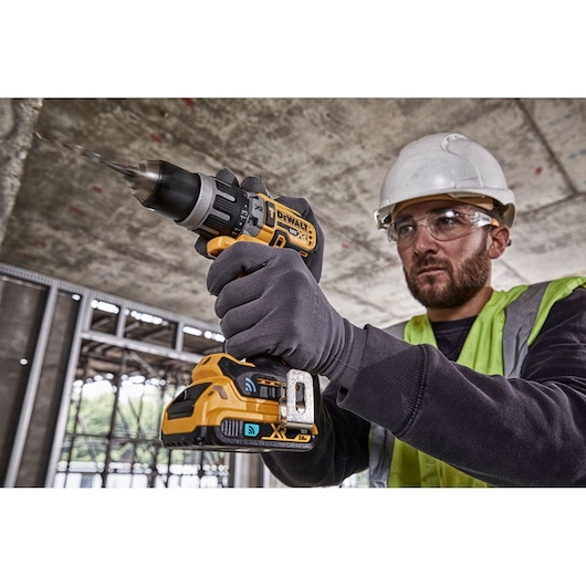 A person wearing a safety helmet, safety vest, and gloves operates a DEWALT cordless drill driver model DCD997NT at a construction site. The drill is being used against a concrete surface.