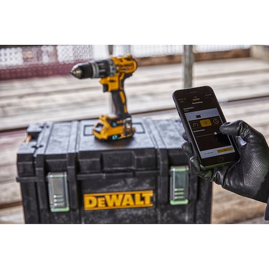 A DEWALT cordless drill is placed on top of a black DEWALT toolbox. In the foreground, a person wearing a black glove is holding a smartphone, which is displaying a DEWALT tool control app interface.