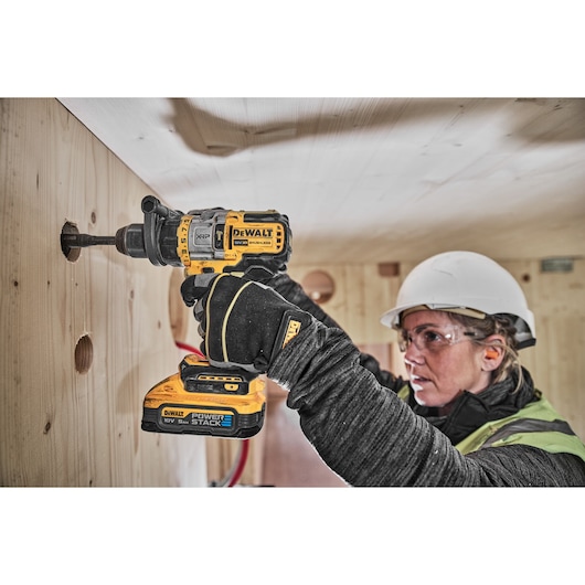 A person wearing safety gear is using a DEWALT DCD999H2T cordless drill to bore a hole into a wooden wall. The drill is fitted with a POWERSTACK battery.