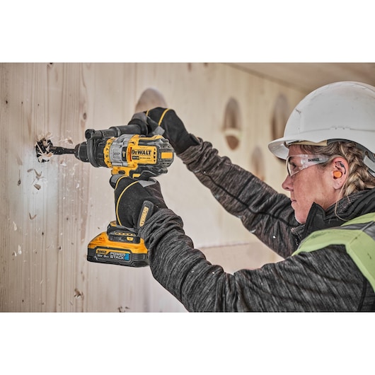 A person wearing safety gear uses a DEWALT DCD999H2T cordless drill to drill a hole in a wooden wall.