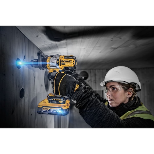 A person wearing safety gear uses a DEWALT DCD999H2T PowerStack cordless drill to bore a hole into a wooden wall. The drill is illuminated, and the person's face is blurred for privacy.