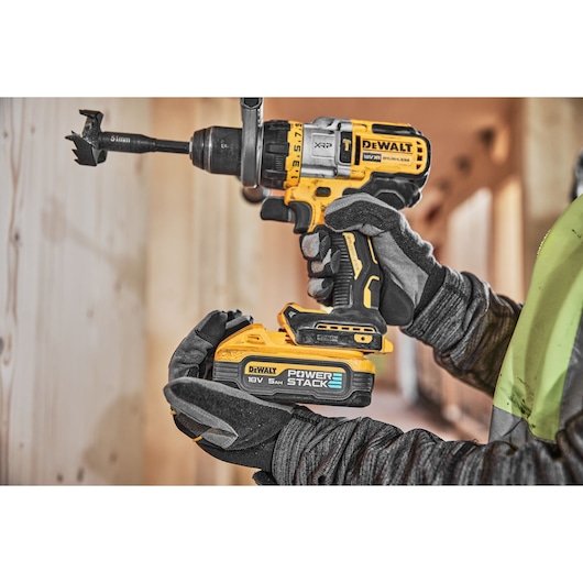 A close-up of a gloved worker holding a DEWALT DCD999H2T cordless drill with a POWERSTACK 18V 5Ah battery, drilling into wood.