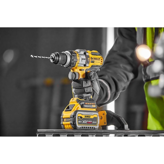 Gloved hand holding a yellow DEWALT cordless drill with battery, shown in a workshop setting.