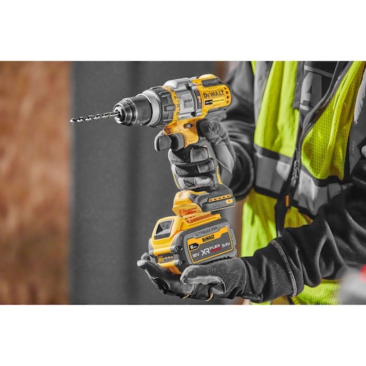 DEWALT cordless drill DCD999NT held by gloved hands at a worksite, with battery attached and drill bit installed.