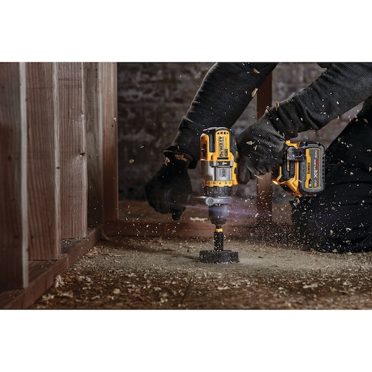 Person uses a DEWALT cordless drill to make a hole in a wooden floor, with wood shavings flying around.