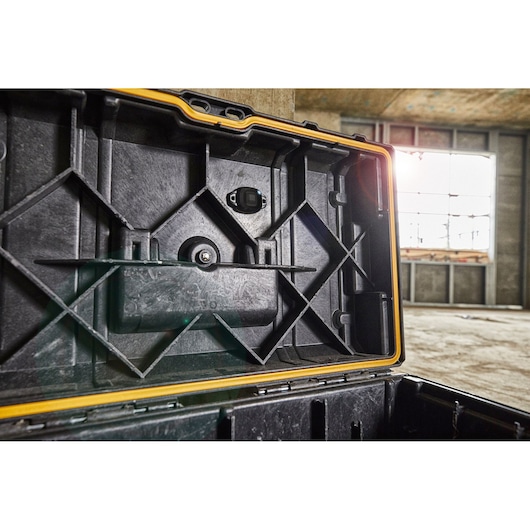 Close-up view of the inside lid of a DEWALT tool storage case, featuring a rugged black plastic construction and yellow trim. The tool case is open in an unfinished industrial building with sunlight coming in through a large window.