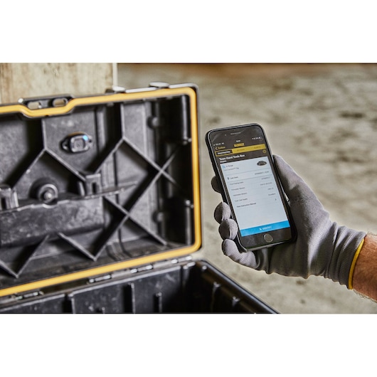 A gloved hand holds a smartphone displaying asset tracking information for a DEWALT tool box next to an open, rugged black and yellow tool case.