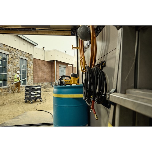 DEWALT device sits atop blue barrel in a worksite, coiled hoses and a worker can be seen outside near a stone building.