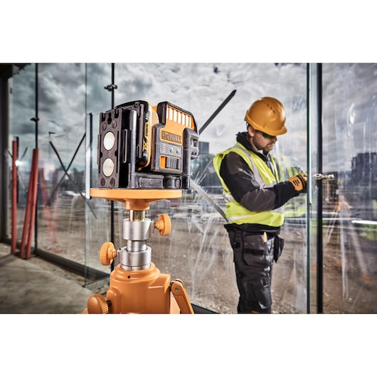 DEWALT green laser level on tripod at construction site, worker installing glass panel in background.