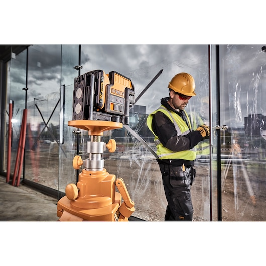 A DEWALT laser level device is mounted on a tripod at a construction site, with a person wearing a safety vest and hard hat working in the background near large glass panels.