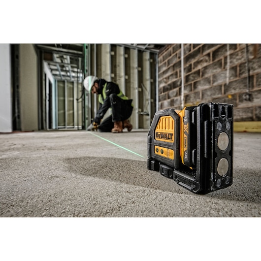 A DEWALT DCE088D1G green beam cross line laser level is placed on the floor at a construction site. In the background, a worker wearing safety equipment is using the laser line for alignment.