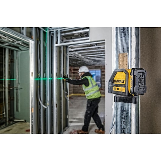 A DEWALT DCE088D1G green laser level mounted on a metal frame at a construction site, projecting a green horizontal laser line. In the background, a construction worker wearing a safety vest and helmet is aligning wall frameworks.