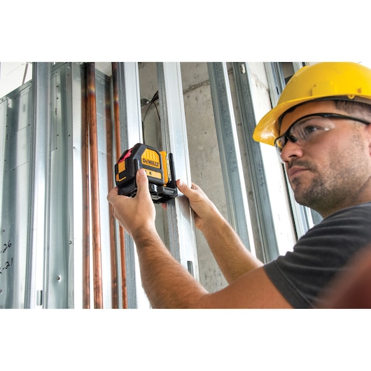 A person wearing a yellow safety helmet uses a DEWALT laser level tool (model DCE088D1R) to align metal studs in a construction setting. The face of the person is blurred for privacy.