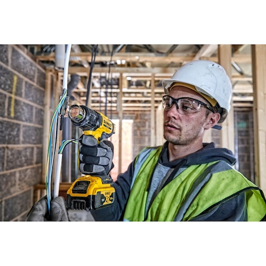 A person wearing safety gear uses a DEWALT DCE530N cordless heat gun to work on electrical wires at a construction site. The individual's face is obscured for privacy.