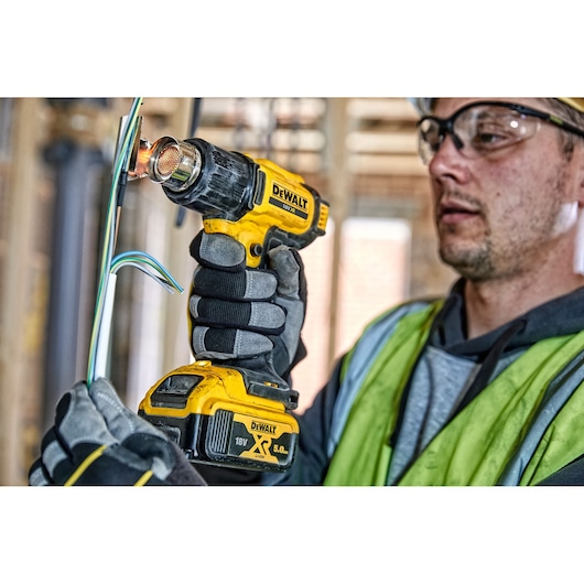A person wearing work gloves and a safety vest uses a DEWALT DCE530N cordless heat gun to apply heat to electrical wires at a construction site. The person's face is blurred for privacy.