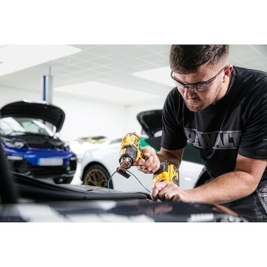 A person using a DEWALT cordless heat gun (model DCE530N) on a car part, with the face blurred for privacy. The individual is wearing a DEWALT-branded shirt and is working in a garage with cars in the background.