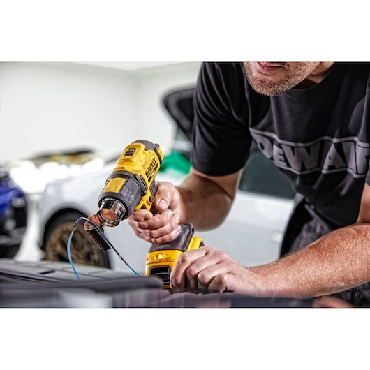 A person uses a DEWALT DCE530N cordless heat gun to work on a surface, with wires and cars visible in the background.