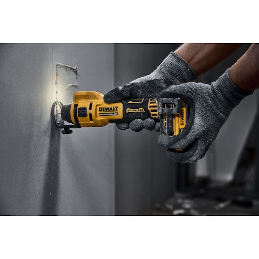 DEWALT cordless rotary tool cutting into drywall, operated by gloved hands.