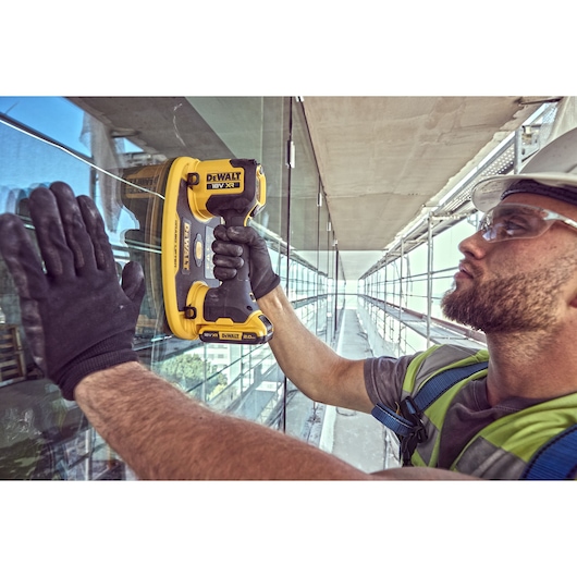 Worker using DEWALT DCE590 tool on glass panel during construction, wearing gloves and safety gear.