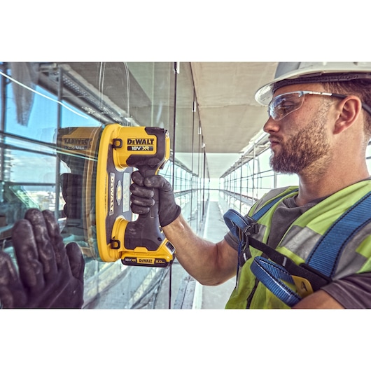 Worker uses DEWALT DCE590 tool to handle glass pane on construction site, wearing safety gear.