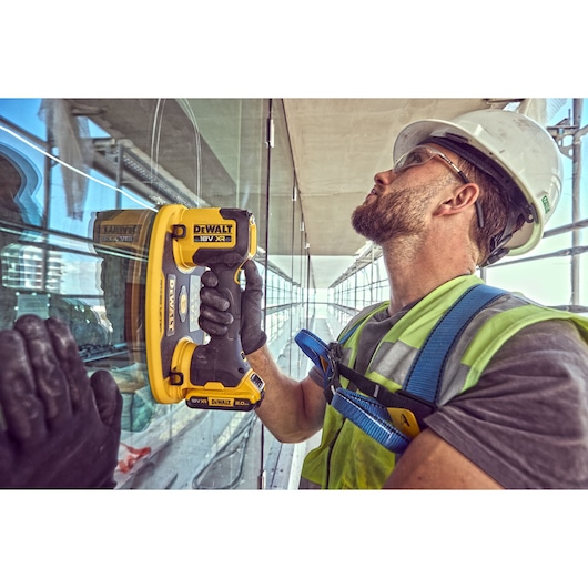Worker uses DEWALT DCE590 tool on glass. Wearing safety gear and helmet at a construction site.