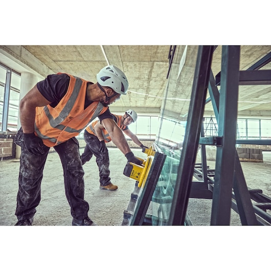 Two workers use DEWALT tools to handle glass on a construction site, wearing helmets and safety vests.