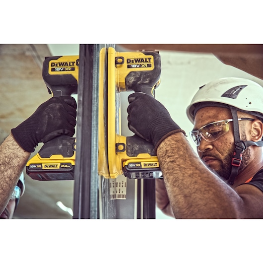Worker using DEWALT DCE590 18V tool to install glass, wearing gloves and helmet on a construction site.